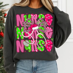 May include: Dark gray sweatshirt with a festive design. The word "NURSE" is displayed in pink and green, with a stethoscope and a bow featuring a leopard print and a Santa hat. The design is decorated with snowflakes and stars.