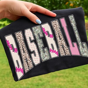 May include: Black t-shirt with the word "BASEBALL" in a bold, stylized font. The letters feature various patterns, including leopard print, camouflage, and pink stripes, with pink baseball stitching details. The shirt is held up by a hand.