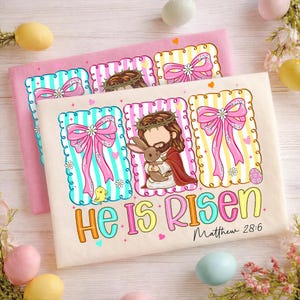 May include: Two Easter-themed decorative prints. One is pink, the other is off-white. Each features a central image of Jesus holding a bunny, surrounded by bows, flowers, and eggs. The text "He Is Risen" is displayed in colourful letters.