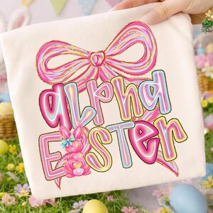 May include: Cream-colored t-shirt featuring the words "Alpha Easter" in a colorful, outlined font. A pink bow and a bunny graphic are above and below the text. The design has a pastel color scheme with a glittery gold outline.