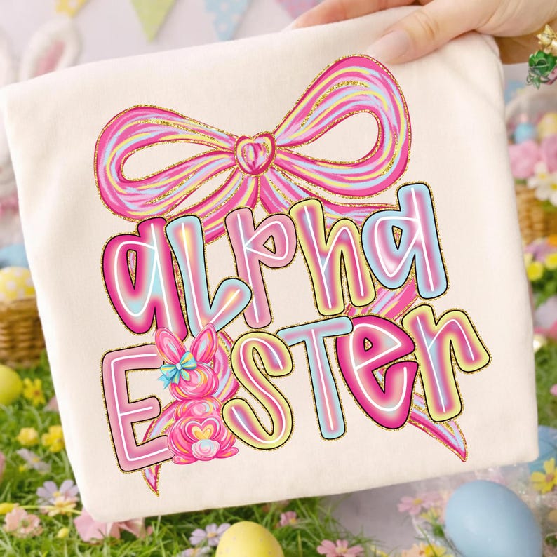 May include: Cream-colored t-shirt featuring the words "Alpha Easter" in a colorful, outlined font. A pink bow and a bunny graphic are above and below the text. The design has a pastel color scheme with a glittery gold outline.