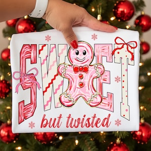 May include: White t-shirt featuring the words "SWEET" and "but twisted" in pink and red. A pink gingerbread man with a red bow tie is in the center. The design has a festive, Christmas theme.