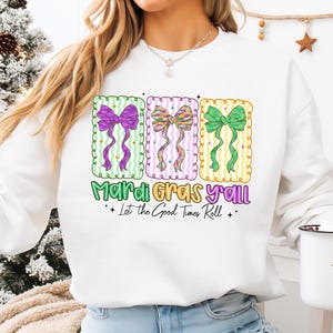 May include: White sweatshirt featuring three colorful bow designs with the text "Mardi Gras y'all" and "Let the Good Times Roll." The bows are purple, multicolored, and green, set against a green, pink, and yellow background.