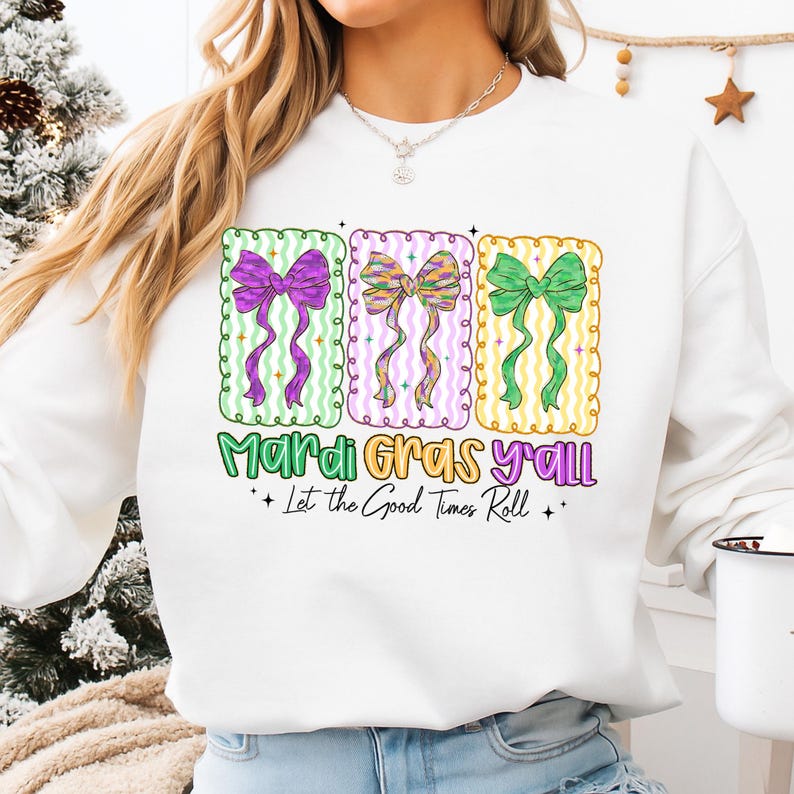May include: White sweatshirt featuring three colorful bow designs with the text "Mardi Gras y'all" and "Let the Good Times Roll." The bows are purple, multicolored, and green, set against a green, pink, and yellow background.