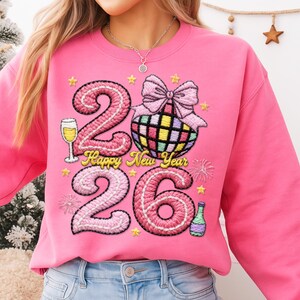 May include: A bright pink sweatshirt with an embroidered design for the New Year. The design includes the numbers "2026", a disco ball, champagne glass, and the words "Happy New Year".