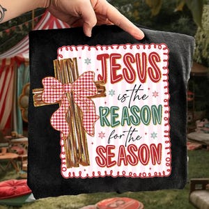 May include: Black t-shirt featuring a decorative cross with a red and white gingham bow. The text "JESUS is the REASON for the SEASON" is displayed in red and green, with snowflake accents, within a decorative border.