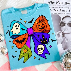 May include: A light blue t-shirt featuring a colorful Halloween bow design. The bow includes a ghost, pumpkin, bats, a skull, and a haunted house. The bow is decorated with glitter and star accents. The shirt is on a pink and white checkered surface.