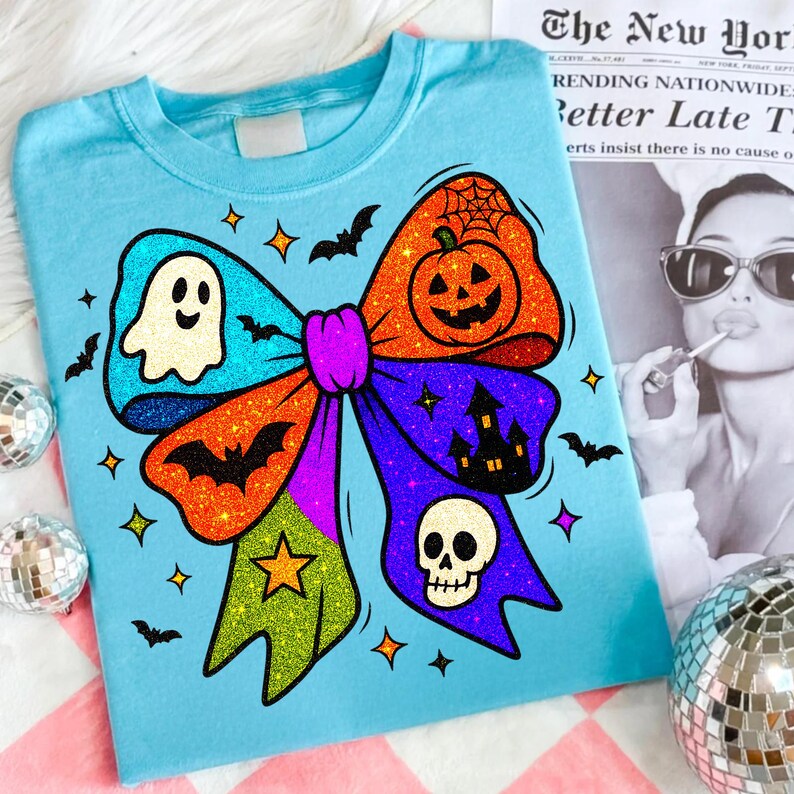 May include: A light blue t-shirt featuring a colorful Halloween bow design. The bow includes a ghost, pumpkin, bats, a skull, and a haunted house. The bow is decorated with glitter and star accents. The shirt is on a pink and white checkered surface.