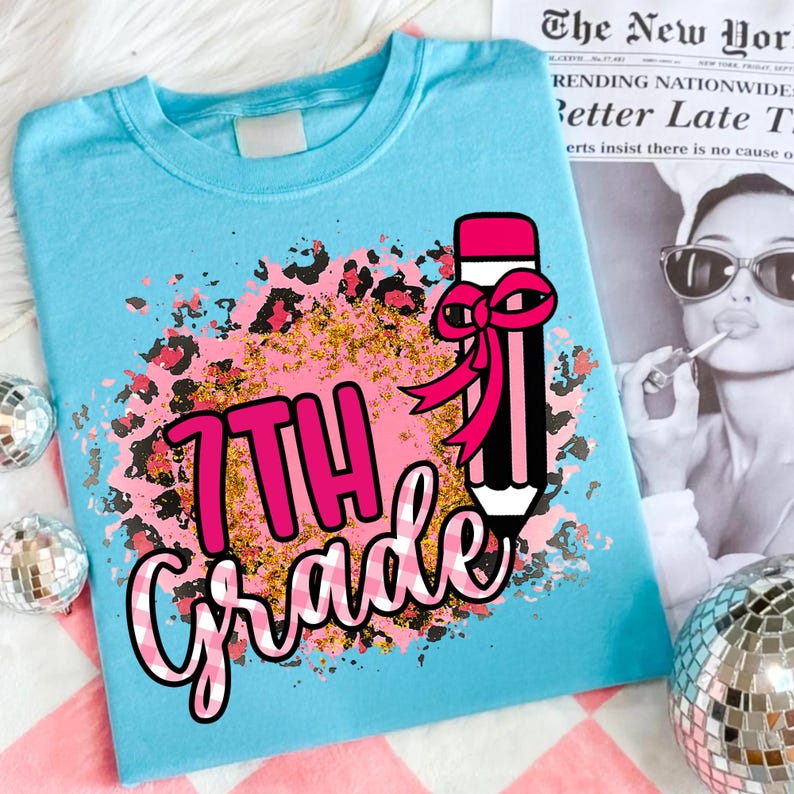 May include: A light blue t-shirt with a colorful graphic design. The design features the words "7th Grade" in pink and white checkered font, a black pencil with a pink bow, and a leopard print and gold glitter background.