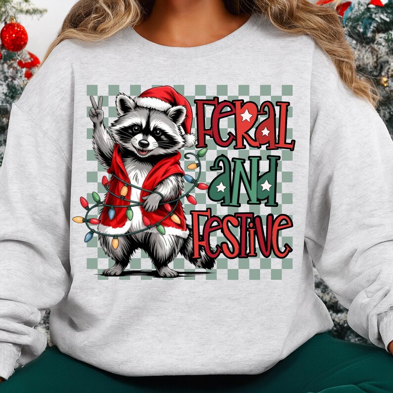 May include: A light gray sweatshirt featuring a raccoon wearing a Santa hat and a red vest adorned with Christmas lights. The raccoon is giving a peace sign. The text "Feral and Festive" is printed on the sweatshirt.