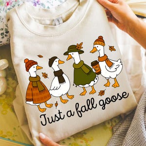 May include: Cream-colored sweatshirt featuring a whimsical illustration of four geese dressed in fall attire. Each goose wears a hat, scarf, and one holds a coffee cup. The text "Just a fall goose" is printed below the geese.