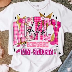 Op de afbeelding: Wit T-shirt met de tekst "MERCY His rewrote MY STORY" in roze, bloemen- en ruitpatronen. Gouden glitter sterren en bloemaccenten omringen de tekst. Het shirt heeft een ronde hals.