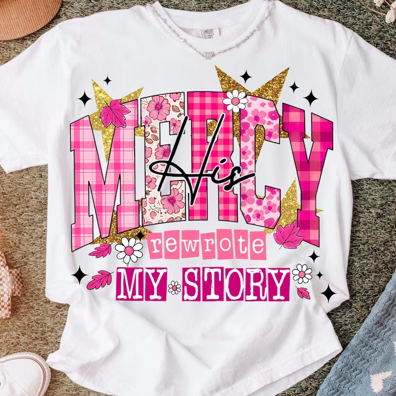 Op de afbeelding: Wit T-shirt met de tekst "MERCY His rewrote MY STORY" in roze, bloemen- en ruitpatronen. Gouden glitter sterren en bloemaccenten omringen de tekst. Het shirt heeft een ronde hals.