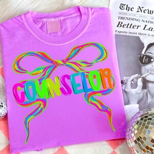 May include: A lilac-colored t-shirt with a colorful bow design and the word "COUNSELOR" in vibrant, multi-colored letters. The design features a rainbow-colored bow and text, creating a playful and eye-catching look. The shirt is a casual wear item.
