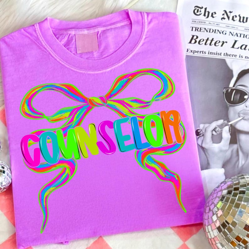 May include: A lilac-colored t-shirt with a colorful bow design and the word "COUNSELOR" in vibrant, multi-colored letters. The design features a rainbow-colored bow and text, creating a playful and eye-catching look. The shirt is a casual wear item.