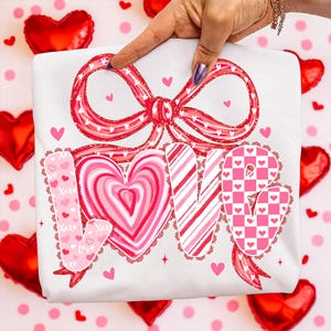 May include: White t-shirt featuring a large red bow design and the word "LOVE" in pink and red. The letters are decorated with hearts, stripes, and a checkerboard pattern. The t-shirt is a Valentine's Day theme.