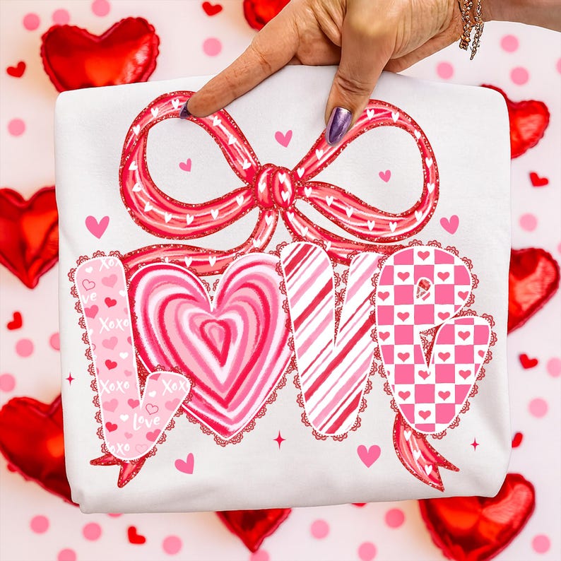 May include: White t-shirt featuring a large red bow design and the word "LOVE" in pink and red. The letters are decorated with hearts, stripes, and a checkerboard pattern. The t-shirt is a Valentine's Day theme.