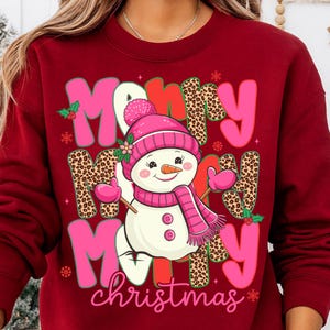 May include: A maroon sweatshirt with a festive design. The design features the words "Merry Christmas" in pink and leopard print, along with a snowman wearing a pink hat, scarf, and mittens. Holly and snowflakes are also included.