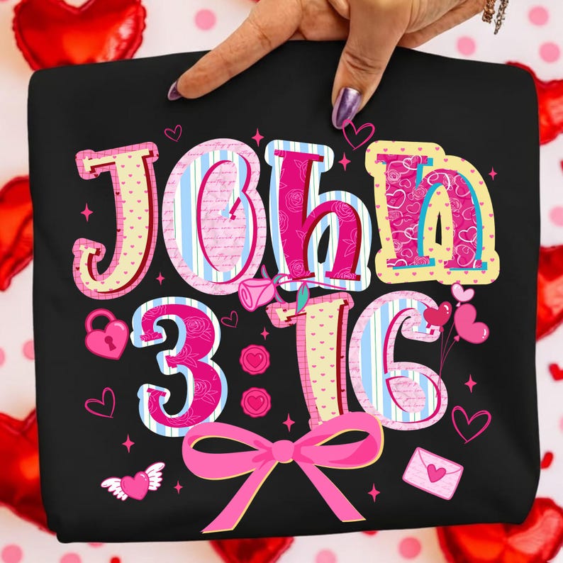 May include: Black t-shirt featuring the text "John 3:16" in pink, yellow, and blue. The design incorporates hearts, a bow, and other decorative elements. The shirt is held by a hand with a purple nail.