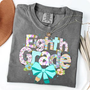 Op de afbeelding: Een grijs Comfort Colors T-shirt met de woorden "Eighth Grade" in een kleurrijk, bloemenontwerp. Het ontwerp bevat een turquoise strik en potlood, met een zilveren ketting over het shirt.