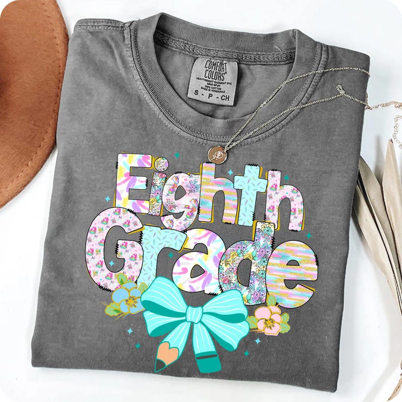 Op de afbeelding: Een grijs Comfort Colors T-shirt met de woorden "Eighth Grade" in een kleurrijk, bloemenontwerp. Het ontwerp bevat een turquoise strik en potlood, met een zilveren ketting over het shirt.