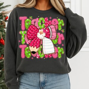 May include: Dark gray sweatshirt with a colorful graphic design. The design features the word "Teacher" in green and pink, a pink bow with a Santa Claus, a pencil, and a notepad. The sweatshirt is a crew neck style.