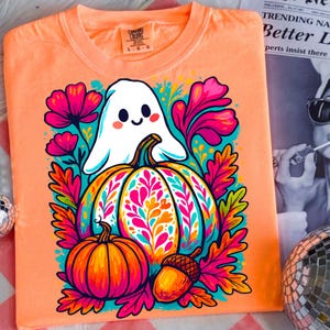 May include: Orange long-sleeve t-shirt featuring a colorful Halloween design. The graphic includes a friendly ghost, a large pumpkin, a smaller pumpkin, and an acorn, surrounded by vibrant flowers and leaves. The shirt has a crew neck and a tag on the upper left.