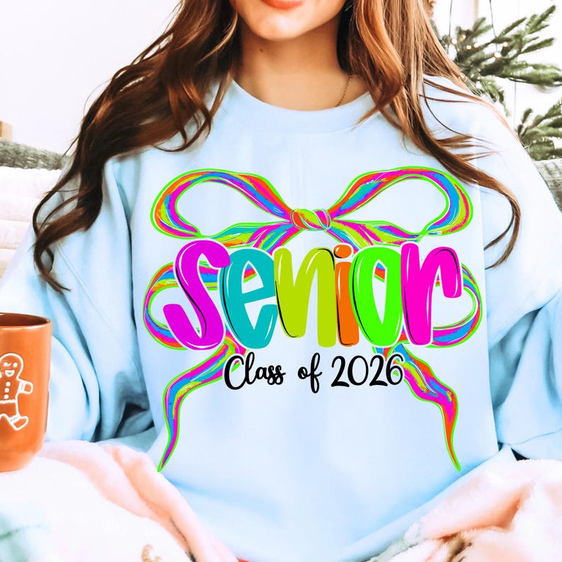May include: Pale blue sweatshirt featuring a colourful bow design and the word "Senior" in pink, green, and orange. The text "Class of 2026" is below. This casual garment is suitable for a high school senior, a popular item for graduation.