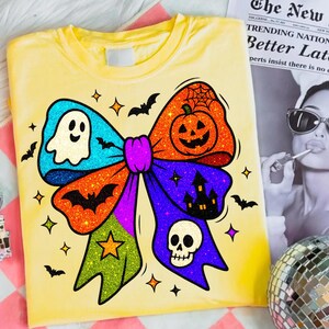 May include: Yellow t-shirt featuring a colorful Halloween bow design. The bow includes a ghost, pumpkin, bat, skull, and haunted house. The design is outlined in black and has glitter accents. Small stars and bats are scattered around the bow.