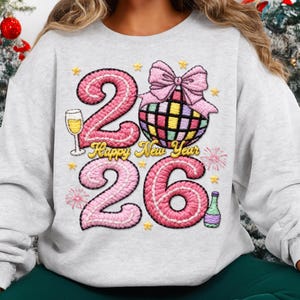 May include: A light gray sweatshirt with a festive New Year's design. The design features the numbers "26" in pink, a disco ball, a champagne glass, and the text "Happy New Year".