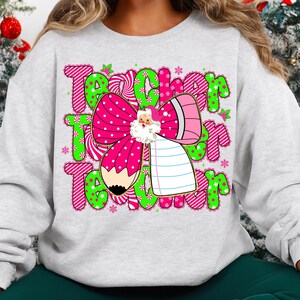 Peut inclure: Un sweat-shirt gris clair avec un motif graphique color&eacute;. Le motif comprend le mot "Teacher" en rose et vert, un n&oelig;ud rose &agrave; pois blancs, un crayon et une illustration du P&egrave;re No&euml;l. Des flocons de neige et du houx compl&egrave;tent le design.