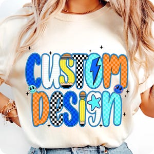 May include: Cream-colored t-shirt with the words "CUSTOM DESIGN" in a colorful, retro font. The design includes a lightning bolt, pencils, stars, and smiley faces. The letters have a checkered pattern.