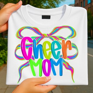 May include: White t-shirt with the words "Cheer Mom" in colorful bubble letters. The text is overlaid on a rainbow-colored bow design. The shirt is held up by a person.