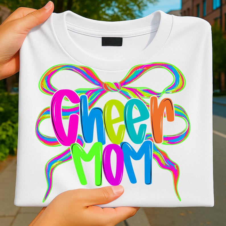May include: White t-shirt with the words "Cheer Mom" in colorful bubble letters. The text is overlaid on a rainbow-colored bow design. The shirt is held up by a person.