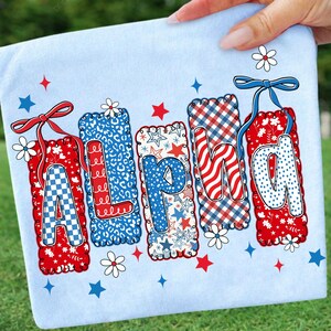 May include: A light blue fabric with the word "ALPHA" in red, white, and blue letters. Each letter has a unique pattern, including checkered, leopard print, stars, and wavy stripes. The letters are decorated with bows, flowers, and stars.