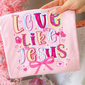 May include: A light pink t-shirt with the words "LOVE like JESUS" in a colorful, whimsical font. The design includes hearts, a bow, and a lock. The shirt is held up by a hand.