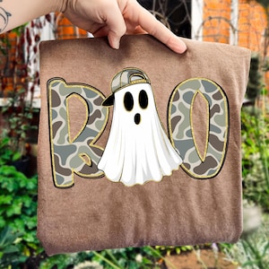 May include: A brown t-shirt featuring the word "BOO" in a camouflage pattern with gold glitter trim. A white ghost wearing a backwards baseball cap is in the center. The ghost has black oval eyes and a black mouth.