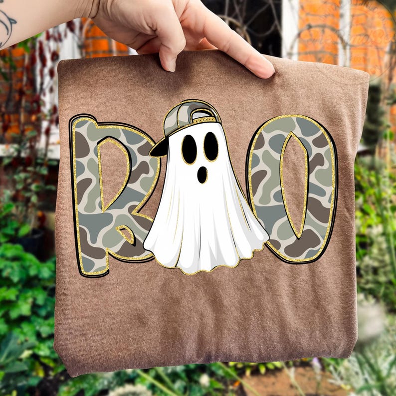 May include: A brown t-shirt featuring the word "BOO" in a camouflage pattern with gold glitter trim. A white ghost wearing a backwards baseball cap is in the center. The ghost has black oval eyes and a black mouth.