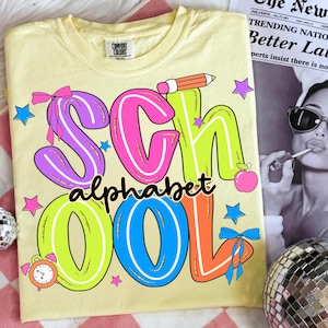 May include: A pale yellow Comfort Colors t-shirt featuring the words "SCHOOL ALPHABET" in vibrant, cartoon lettering. The design incorporates a pencil, apple, stars, and bows, ideal for back-to-school or teacher presents.