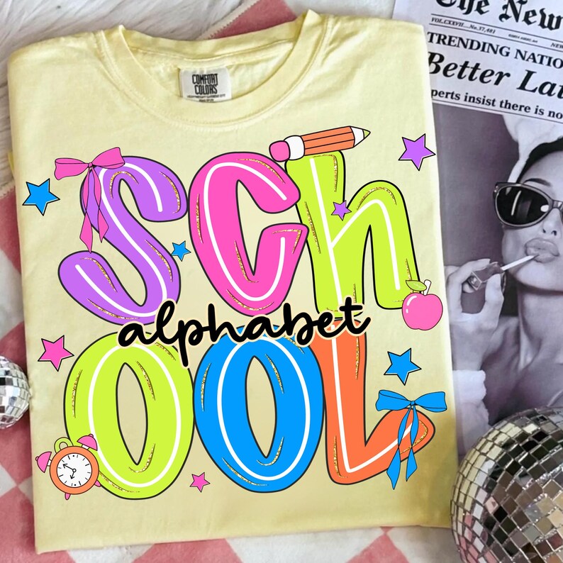 May include: A pale yellow Comfort Colors t-shirt featuring the words "SCHOOL ALPHABET" in vibrant, cartoon lettering. The design incorporates a pencil, apple, stars, and bows, ideal for back-to-school or teacher presents.