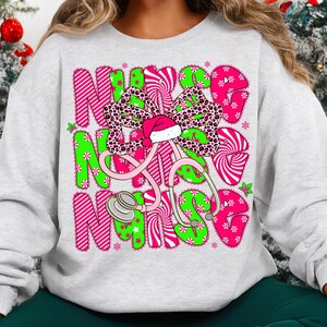 May include: A light gray sweatshirt with a colorful graphic design. The design features the word "NURSE" in pink and green letters, a stethoscope, and a bow with a Santa hat. The bow has a leopard print pattern.