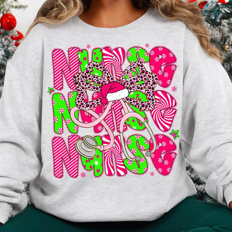 May include: A light gray sweatshirt with a colorful graphic design. The design features the word "NURSE" in pink and green letters, a stethoscope, and a bow with a Santa hat. The bow has a leopard print pattern.