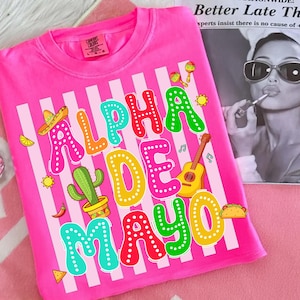 May include: A bright pink t-shirt with the words "ALPHA DE MAYO" in colorful, dotted letters. The design includes a sombrero, cactus, guitar, maracas, and a taco, all set against vertical white stripes.