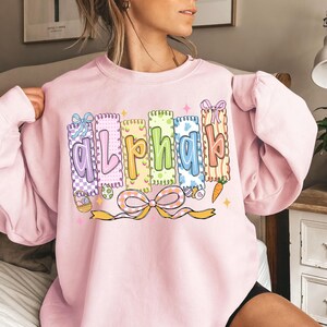 May include: A light pink sweatshirt with the word "Alpha" in colorful, decorative letters. Each letter is designed with a unique pattern and color, including pastel shades and Easter-themed elements like eggs and carrots. A large bow is at the bottom.