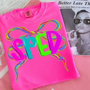 May include: Bright pink t-shirt with the word "SPED" in large, colorful letters. The letters are outlined in black and have a 3D effect. A large, multi-colored bow design is above the letters. The shirt has a tag that says "Comfort Colors".