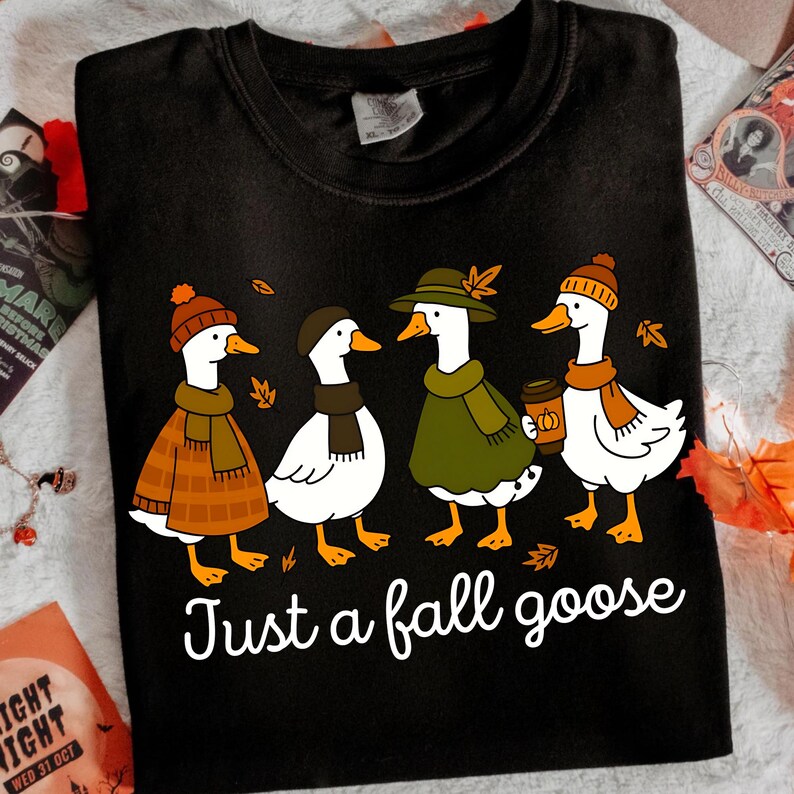 May include: Black t-shirt featuring four cartoon geese dressed in fall attire. The geese wear hats, scarves, and coats, with one holding a coffee cup. The text "Just a fall goose" is printed below the geese.