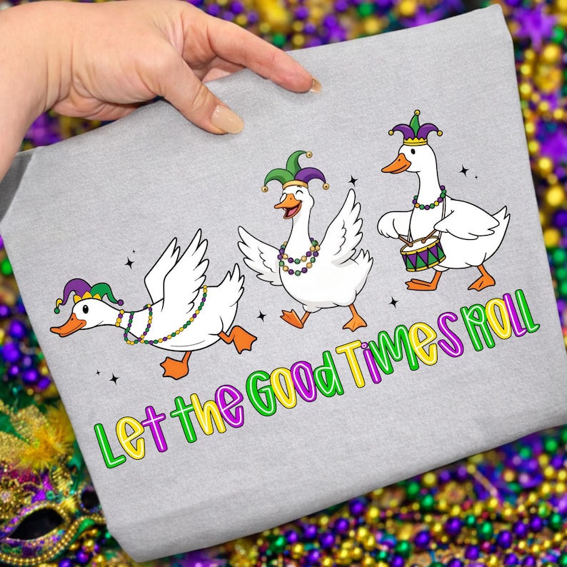 May include: A light gray t-shirt featuring three cartoon geese in Mardi Gras attire. The geese are white with orange feet and beaks, wearing jester hats and beaded necklaces. The text "Let the Good Times Roll" is below the geese.