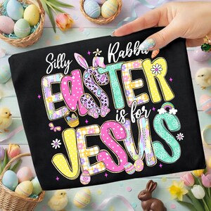 May include: Black t-shirt with the text "Silly Rabbit Easter is for Jesus" in colorful, patterned letters. The design includes a bunny, eggs, and flowers, perfect for Easter celebrations.