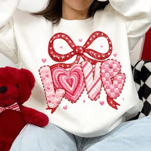 May include: White sweatshirt with a red bow and the word "LOVE" in pink, heart-shaped letters. The "O" is a heart. Small pink hearts are scattered around the design. A red teddy bear is visible on the left.