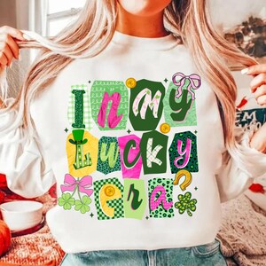 May include: White sweatshirt with a colorful graphic that says "I'm My Lucky Era." The design features green, pink, and gold accents, including shamrocks, a horseshoe, and a leprechaun hat. The text is in a playful, blocky font.
